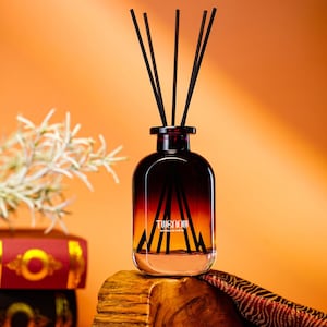 May include: A decorative glass diffuser bottle with black reeds. The bottle has a gradient color scheme, transitioning from dark brown to orange. The text "TWENOW" is visible on the front. The diffuser sits on a wooden block.