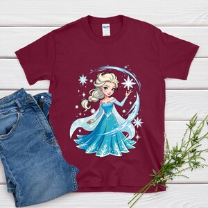 May include: A maroon t-shirt featuring a cartoon illustration of a princess in a blue dress with a snowflake design. The image includes a pair of blue jeans and a sprig of greenery.