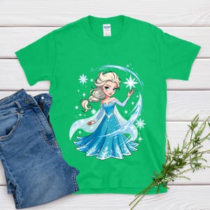 May include: A green t-shirt featuring a cartoon illustration of a princess in a blue dress with white accents and snowflakes. The princess has blonde hair and is surrounded by a blue and white design. Blue jeans are next to the shirt.