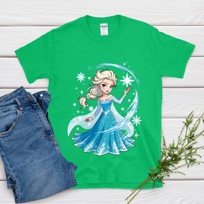 May include: A green t-shirt featuring a cartoon illustration of a princess in a blue dress with white accents and snowflakes. The princess has blonde hair and is surrounded by a blue and white design. Blue jeans are next to the shirt.