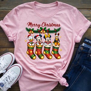 May include: A light pink t-shirt featuring Disney characters in Christmas stockings. The text "Merry Christmas" is above the characters. Donald Duck, Mickey Mouse, Minnie Mouse, and Pluto are in the stockings. White sneakers and blue jeans are also in the image.