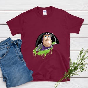 May include: Burgundy t-shirt featuring a graphic of Buzz Lightyear from Toy Story. The design shows Buzz in his space suit, with a green splash effect. A pair of blue jeans is next to the shirt.