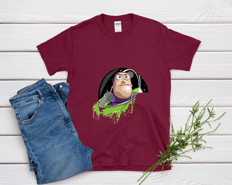 May include: Burgundy t-shirt featuring a graphic of Buzz Lightyear from Toy Story. The design shows Buzz in his space suit, with a green splash effect. A pair of blue jeans is next to the shirt.