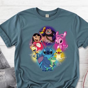 Op de afbeelding: Een tealkleurig T-shirt met een kleurrijke afbeelding van personages uit de animatiefilm Lilo & Stitch. Het ontwerp bevat Stitch, Lilo en andere personages met hun namen in cursief. Het shirt wordt getoond met een spijkerbroek en witte sneakers.