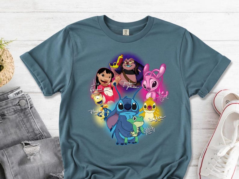 Op de afbeelding: Een tealkleurig T-shirt met een kleurrijke afbeelding van personages uit de animatiefilm Lilo & Stitch. Het ontwerp bevat Stitch, Lilo en andere personages met hun namen in cursief. Het shirt wordt getoond met een spijkerbroek en witte sneakers.
