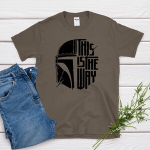 May include: A brown t-shirt with the text "THIS IS THE WAY" in black, along with a black helmet graphic. The shirt is paired with blue jeans. The design is likely inspired by a popular science fiction franchise.