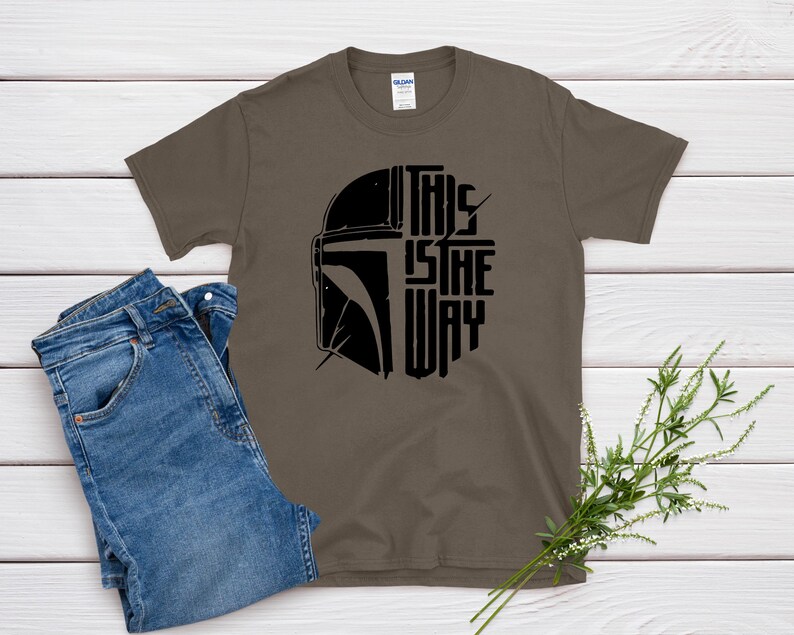 May include: A brown t-shirt with the text "THIS IS THE WAY" in black, along with a black helmet graphic. The shirt is paired with blue jeans. The design is likely inspired by a popular science fiction franchise.