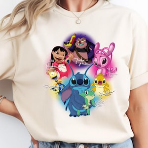 Op de afbeelding: Beige T-shirt met een kleurrijke afbeelding van personages uit de animatiefilm Lilo & Stitch. Het ontwerp bevat Stitch, Lilo en andere personages, met hun namen in handschrift. Het shirt heeft korte mouwen en een ronde hals.