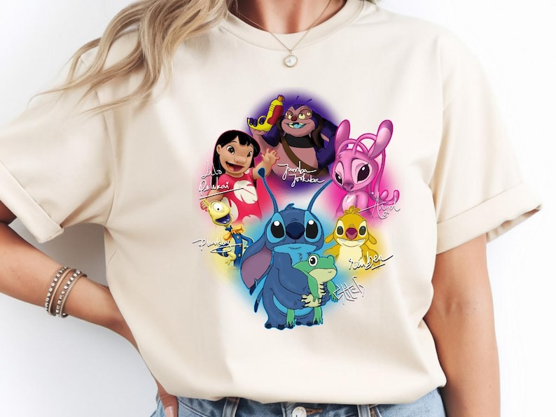 Op de afbeelding: Beige T-shirt met een kleurrijke afbeelding van personages uit de animatiefilm Lilo & Stitch. Het ontwerp bevat Stitch, Lilo en andere personages, met hun namen in handschrift. Het shirt heeft korte mouwen en een ronde hals.