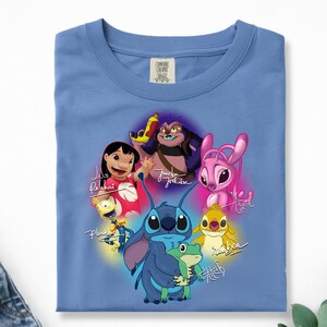 Op de afbeelding: Een lichtblauw T-shirt met een kleurrijke afbeelding van personages uit de animatiefilm Lilo & Stitch. Het ontwerp bevat Stitch, Lilo en andere personages met hun namen in cursief. Het shirt is gemaakt van een zacht materiaal.