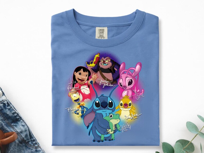 Op de afbeelding: Een lichtblauw T-shirt met een kleurrijke afbeelding van personages uit de animatiefilm Lilo & Stitch. Het ontwerp bevat Stitch, Lilo en andere personages met hun namen in cursief. Het shirt is gemaakt van een zacht materiaal.