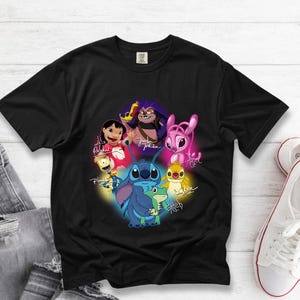 Op de afbeelding: Zwart T-shirt met een kleurrijke afbeelding van personages uit de animatiefilm Lilo & Stitch, waaronder Stitch en Lilo, met hun handtekeningen. Het ontwerp is geplaatst tegen een zwarte achtergrond. Het T-shirt ligt op een wit houten oppervlak.