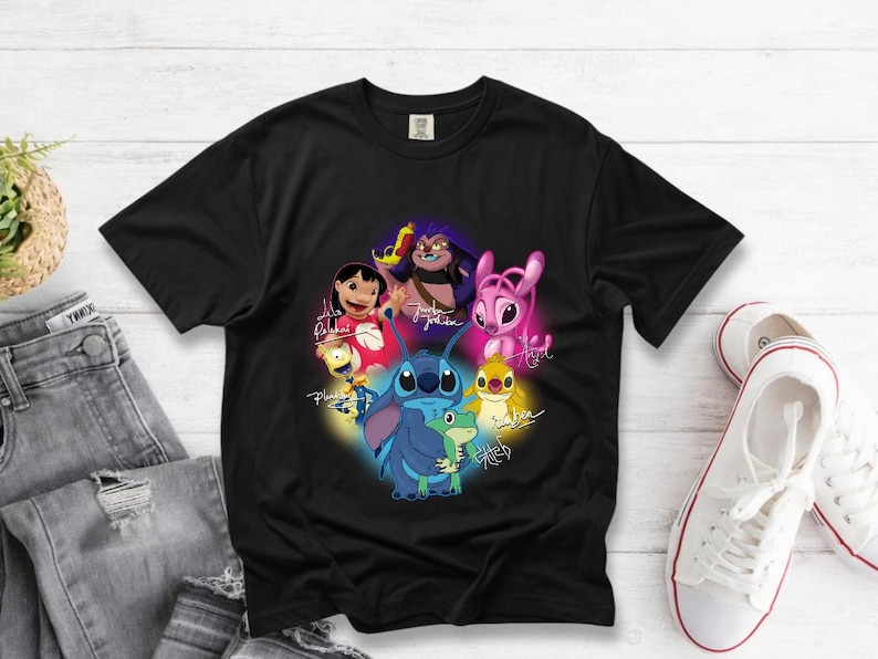 Op de afbeelding: Zwart T-shirt met een kleurrijke afbeelding van personages uit de animatiefilm Lilo & Stitch, waaronder Stitch en Lilo, met hun handtekeningen. Het ontwerp is geplaatst tegen een zwarte achtergrond. Het T-shirt ligt op een wit houten oppervlak.