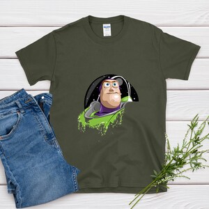 May include: Olive green t-shirt featuring a graphic of Buzz Lightyear from Toy Story. The design shows Buzz in his space suit with a green and purple color scheme. A pair of blue jeans is next to the shirt.
