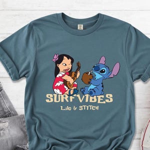 Puede incluir: Camiseta azul verdoso con un dise&ntilde;o de Lilo y Stitch. Lilo toca el ukelele y Stitch bebe de un coco. El texto "SURF VIBES" y "Lilo & Stitch" est&aacute; impreso en color beige.