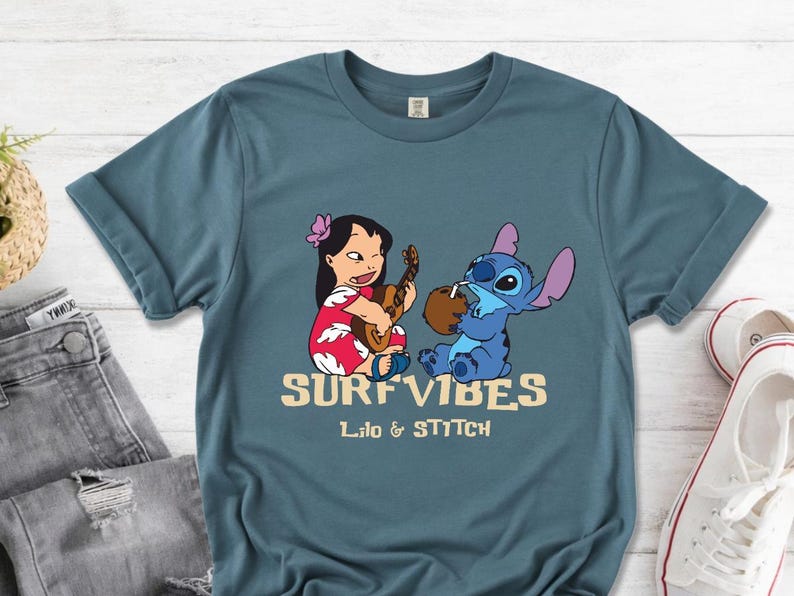 Puede incluir: Camiseta azul verdoso con un dise&ntilde;o de Lilo y Stitch. Lilo toca el ukelele y Stitch bebe de un coco. El texto "SURF VIBES" y "Lilo & Stitch" est&aacute; impreso en color beige.