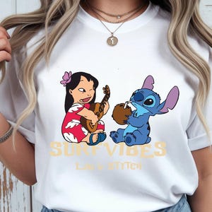 Puede incluir: Camiseta blanca con un dise&ntilde;o de Lilo y Stitch. Lilo toca el ukelele y Stitch bebe de un coco. Las palabras "SURF VIBES" y "LILO & STITCH" est&aacute;n impresas en amarillo.