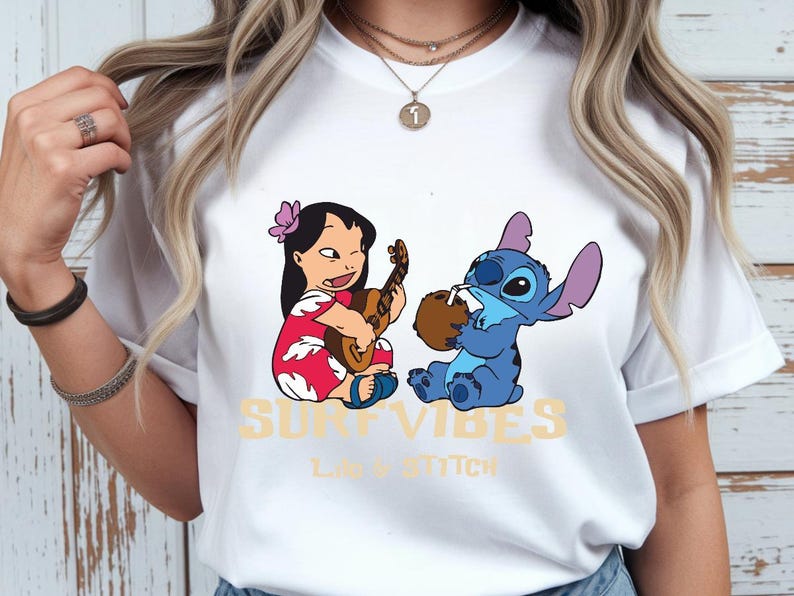 Puede incluir: Camiseta blanca con un dise&ntilde;o de Lilo y Stitch. Lilo toca el ukelele y Stitch bebe de un coco. Las palabras "SURF VIBES" y "LILO & STITCH" est&aacute;n impresas en amarillo.