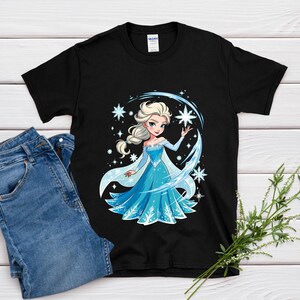 May include: Black t-shirt featuring a cartoon illustration of a princess in a blue dress with snowflakes and a star. The princess has blonde hair and is surrounded by a blue arc and stars. A pair of blue jeans is next to the shirt.