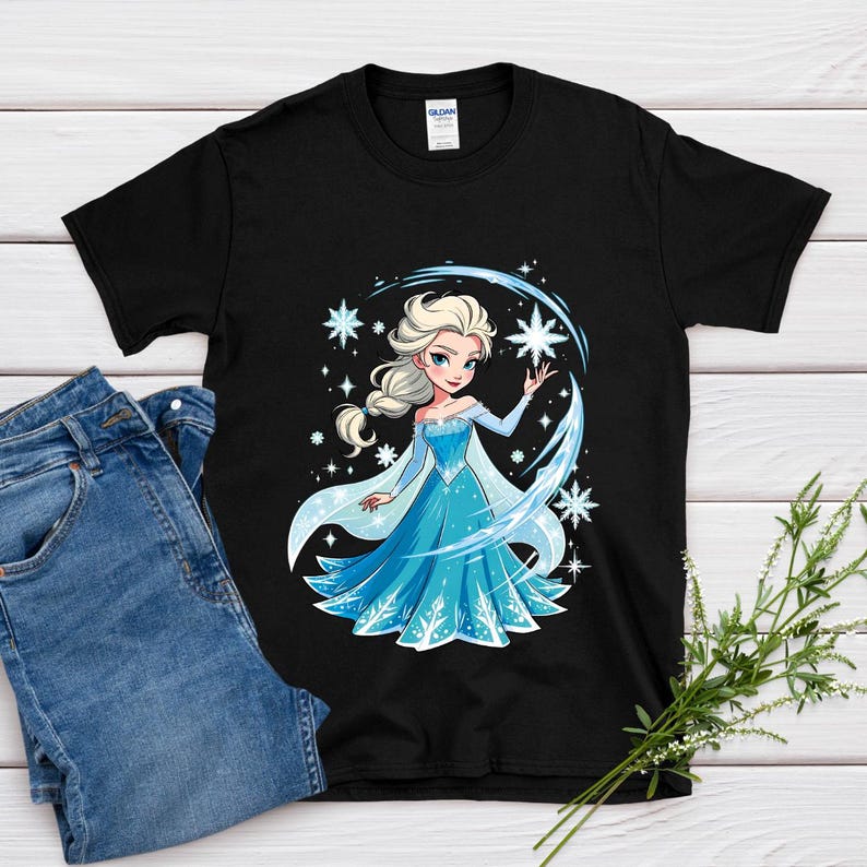 May include: Black t-shirt featuring a cartoon illustration of a princess in a blue dress with snowflakes and a star. The princess has blonde hair and is surrounded by a blue arc and stars. A pair of blue jeans is next to the shirt.