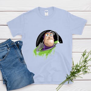 May include: A light blue t-shirt featuring a graphic of Buzz Lightyear from Toy Story. The design shows Buzz in his space suit against a starry background. A pair of blue jeans is next to the shirt.