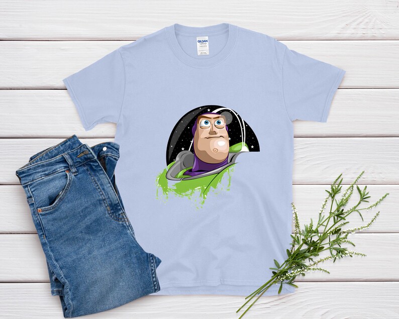 May include: A light blue t-shirt featuring a graphic of Buzz Lightyear from Toy Story. The design shows Buzz in his space suit against a starry background. A pair of blue jeans is next to the shirt.