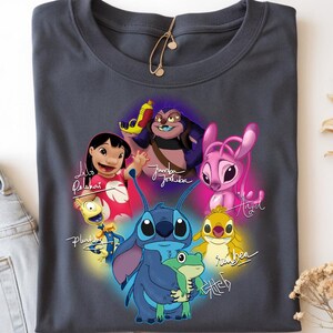 Op de afbeelding: Donkergrijs t-shirt met een kleurrijke afbeelding van personages uit de animatiefilm Lilo & Stitch. Het ontwerp bevat Stitch, Lilo, Angel en andere personages, met hun namen in cursief geschreven. Het shirt is opgevouwen.