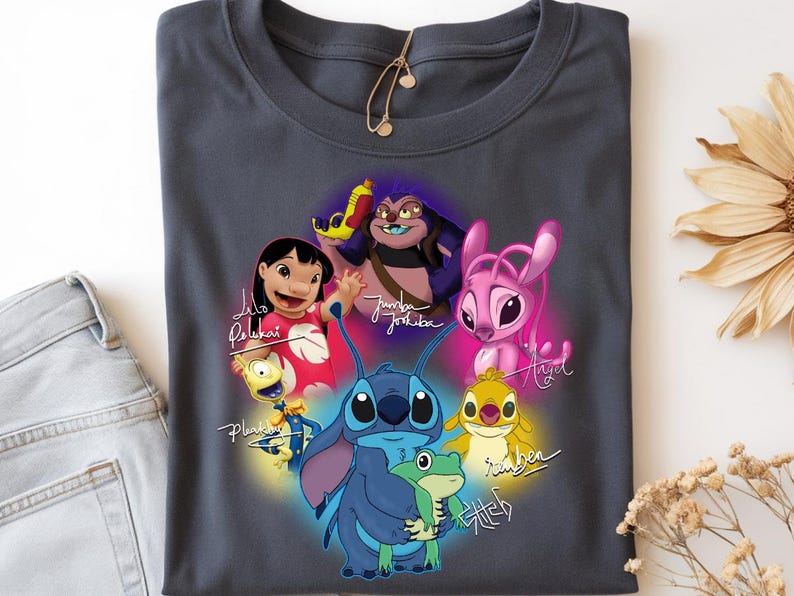 Op de afbeelding: Donkergrijs t-shirt met een kleurrijke afbeelding van personages uit de animatiefilm Lilo & Stitch. Het ontwerp bevat Stitch, Lilo, Angel en andere personages, met hun namen in cursief geschreven. Het shirt is opgevouwen.