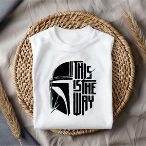May include: White t-shirt with a black graphic of a helmet and the text "THIS IS THE WAY". The shirt is displayed on a woven basket. The design is a bold, graphic statement.