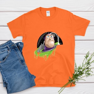 May include: Orange t-shirt featuring a graphic of Buzz Lightyear in a spaceship against a black and starry background. The shirt is paired with blue jeans on a white wooden surface.