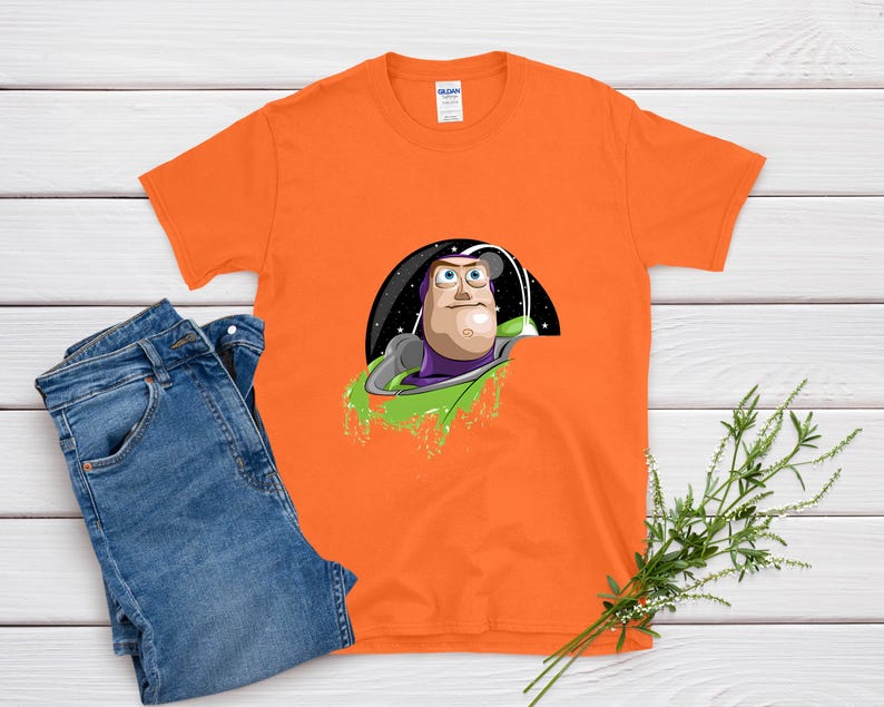 May include: Orange t-shirt featuring a graphic of Buzz Lightyear in a spaceship against a black and starry background. The shirt is paired with blue jeans on a white wooden surface.