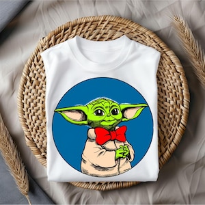 May include: White t-shirt featuring a cartoon character with green skin, large ears, and a red bow tie, set against a blue circle. The character is smiling and wearing a tan robe. The shirt is folded on a woven basket.