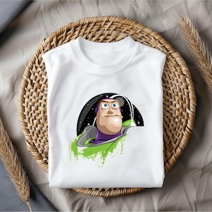 May include: White t-shirt featuring a cartoon illustration of Buzz Lightyear from Toy Story. The graphic shows Buzz in his space suit with a black and starry background. The shirt is displayed on a woven basket.