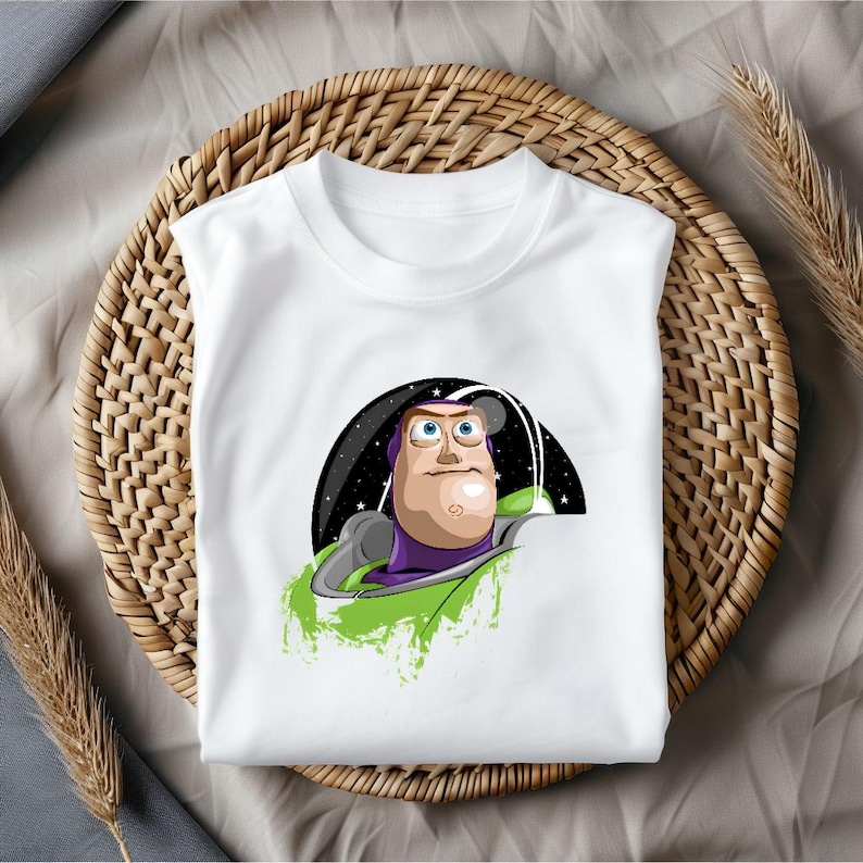 May include: White t-shirt featuring a cartoon illustration of Buzz Lightyear from Toy Story. The graphic shows Buzz in his space suit with a black and starry background. The shirt is displayed on a woven basket.