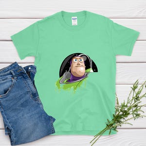 May include: A light green t-shirt featuring a graphic of Buzz Lightyear from Toy Story. The cartoon character is set against a black and starry background. A pair of blue jeans is next to the t-shirt. A sprig of greenery is in the bottom right corner.