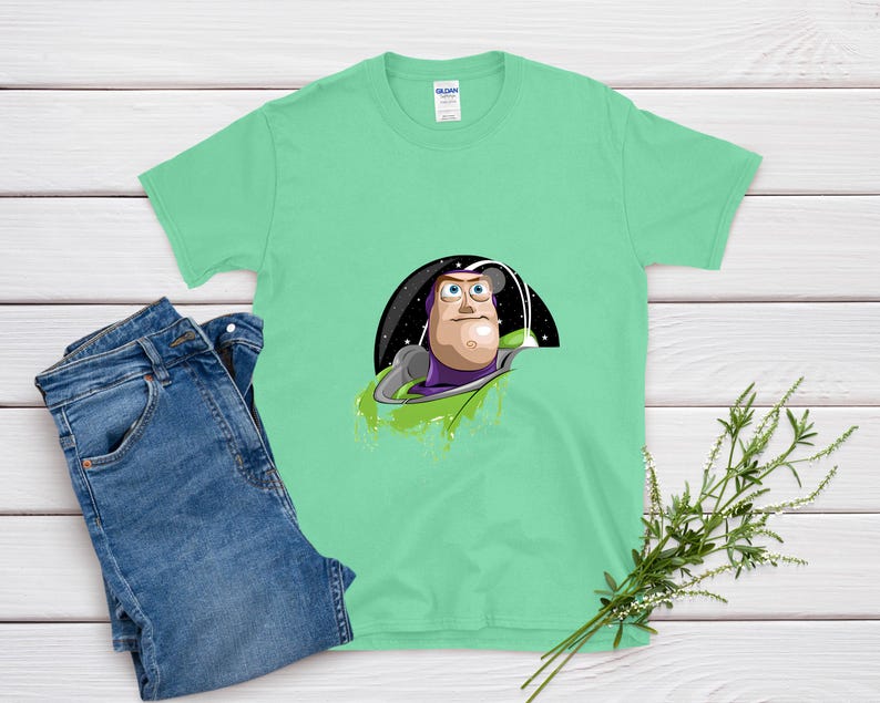 May include: A light green t-shirt featuring a graphic of Buzz Lightyear from Toy Story. The cartoon character is set against a black and starry background. A pair of blue jeans is next to the t-shirt. A sprig of greenery is in the bottom right corner.