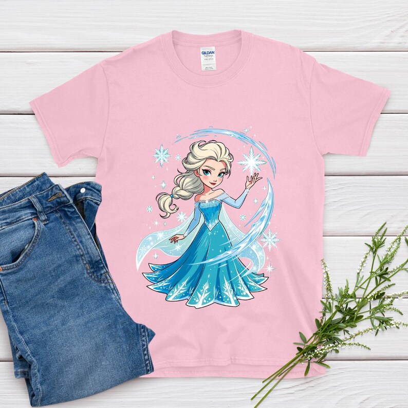 May include: A light pink t-shirt featuring a cartoon illustration of a princess in a blue dress with snowflakes. The princess has blonde hair and is surrounded by a blue arc and stars. A pair of blue jeans and greenery are also in the image.