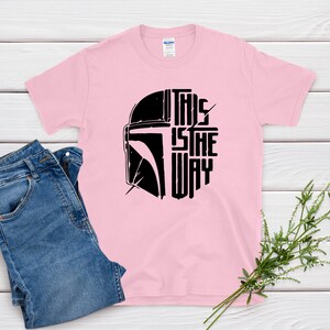 May include: A light pink t-shirt with a black graphic of a helmet and the text "THIS IS THE WAY." Blue denim jeans are folded next to the shirt. A sprig of greenery is placed near the shirt.