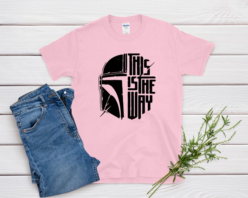 May include: A light pink t-shirt with a black graphic of a helmet and the text "THIS IS THE WAY." Blue denim jeans are folded next to the shirt. A sprig of greenery is placed near the shirt.