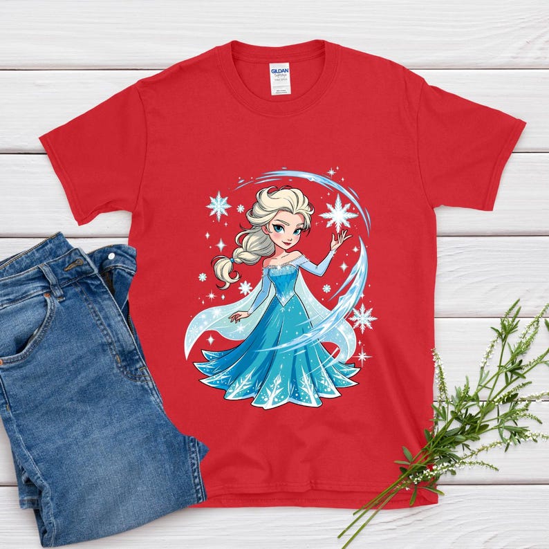 May include: Red t-shirt featuring a cartoon illustration of a princess in a blue dress with white accents and snowflakes. The princess has blonde hair and is surrounded by stars and a blue arc. Blue jeans are next to the shirt.