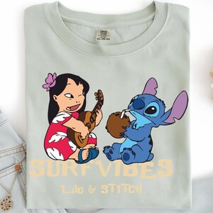 Puede incluir: Camiseta verde claro con una ilustraci&oacute;n de dibujos animados de Lilo tocando el ukelele y Stitch bebiendo de un coco. El texto "SURF VIBES" y "Lilo & Stitch" est&aacute;n impresos en color naranja claro.