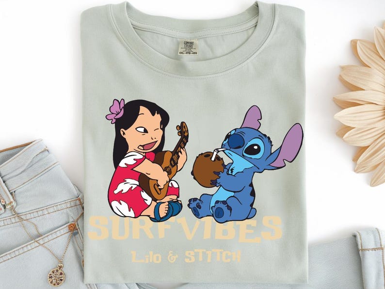 Puede incluir: Camiseta verde claro con una ilustraci&oacute;n de dibujos animados de Lilo tocando el ukelele y Stitch bebiendo de un coco. El texto "SURF VIBES" y "Lilo & Stitch" est&aacute;n impresos en color naranja claro.