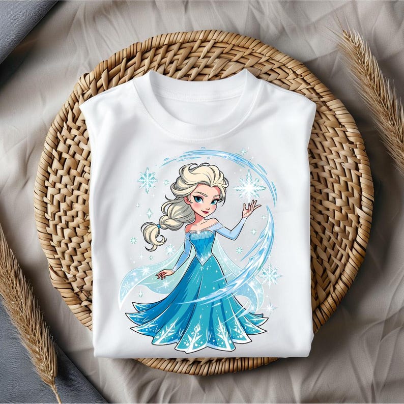May include: White t-shirt featuring a cartoon illustration of a blonde woman in a blue dress. The design includes snowflakes and a blue circular frame. The shirt is displayed on a woven basket.