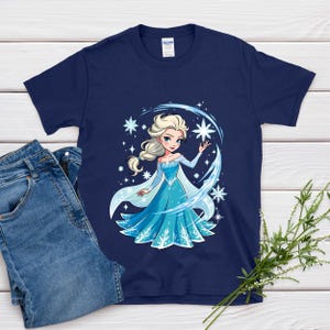 May include: Navy blue t-shirt featuring a cartoon illustration of a princess in a blue dress with snowflakes. The princess has blonde hair and is surrounded by stars and ice-like accents. A pair of blue jeans is next to the shirt.