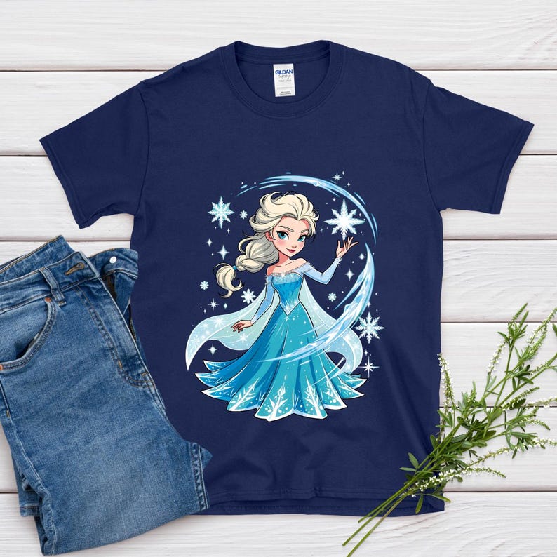 May include: Navy blue t-shirt featuring a cartoon illustration of a princess in a blue dress with snowflakes. The princess has blonde hair and is surrounded by stars and ice-like accents. A pair of blue jeans is next to the shirt.