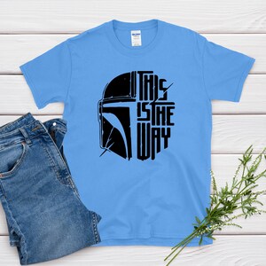 May include: A light blue t-shirt featuring a black graphic of a helmet and the text "THIS IS THE WAY". The shirt is paired with blue jeans. The shirt is made of a soft material.