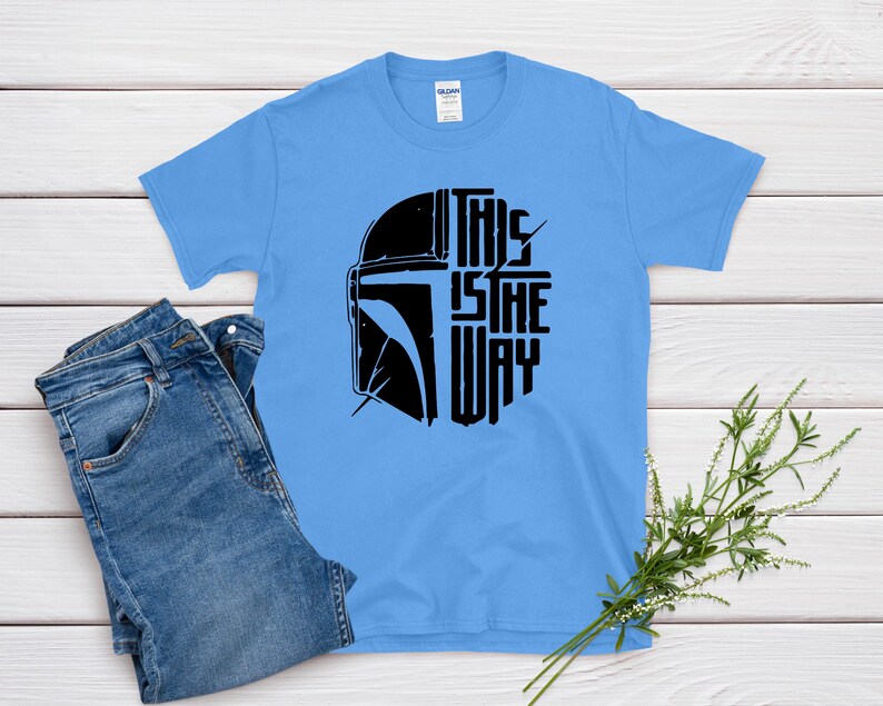 May include: A light blue t-shirt featuring a black graphic of a helmet and the text "THIS IS THE WAY". The shirt is paired with blue jeans. The shirt is made of a soft material.