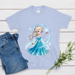 May include: A light blue t-shirt featuring a cartoon illustration of a princess in a blue dress with a snowflake design. The princess has blonde hair and is surrounded by snowflakes. A pair of blue jeans is next to the shirt.