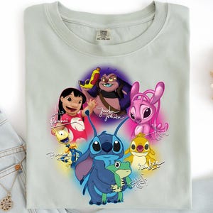 Op de afbeelding: Een lichtgroen t-shirt met een kleurrijke afbeelding van personages uit de animatiefilm Lilo & Stitch. Het ontwerp bevat Stitch, Lilo en andere personages, met hun namen in een gestileerd lettertype. Het t-shirt is opgevouwen.
