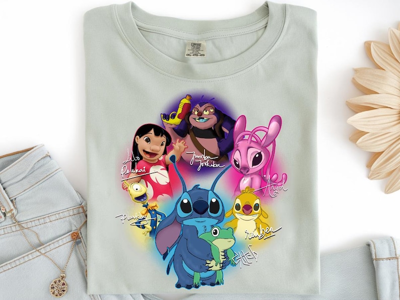 Op de afbeelding: Een lichtgroen t-shirt met een kleurrijke afbeelding van personages uit de animatiefilm Lilo & Stitch. Het ontwerp bevat Stitch, Lilo en andere personages, met hun namen in een gestileerd lettertype. Het t-shirt is opgevouwen.